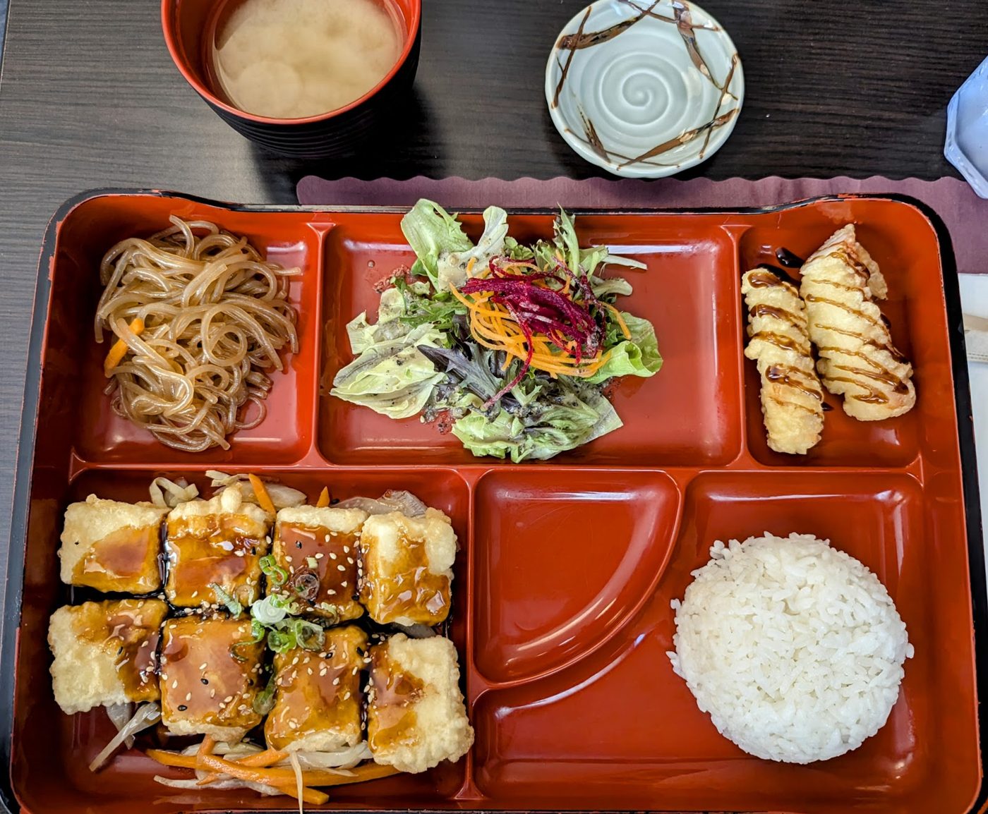 A bento box with rice, salad, noodles and grilled chicken on a red tray
